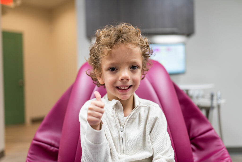 Curly haired boy sitting in dental exam room giving thumbs up to camera